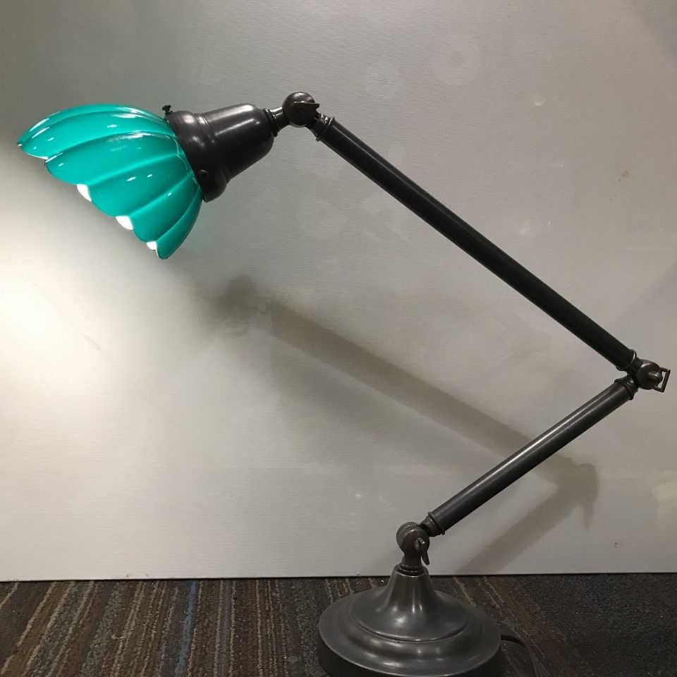 Articulating desk lamp with teal, fluted glass shade, black arm and base, set against a gray wall.