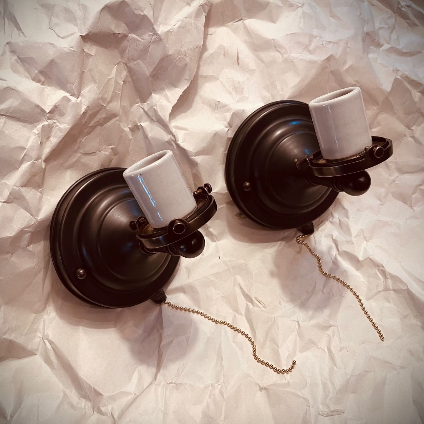 Two bronze-toned wall sconces with ceramic bulb holders and pull chains on crumpled paper.