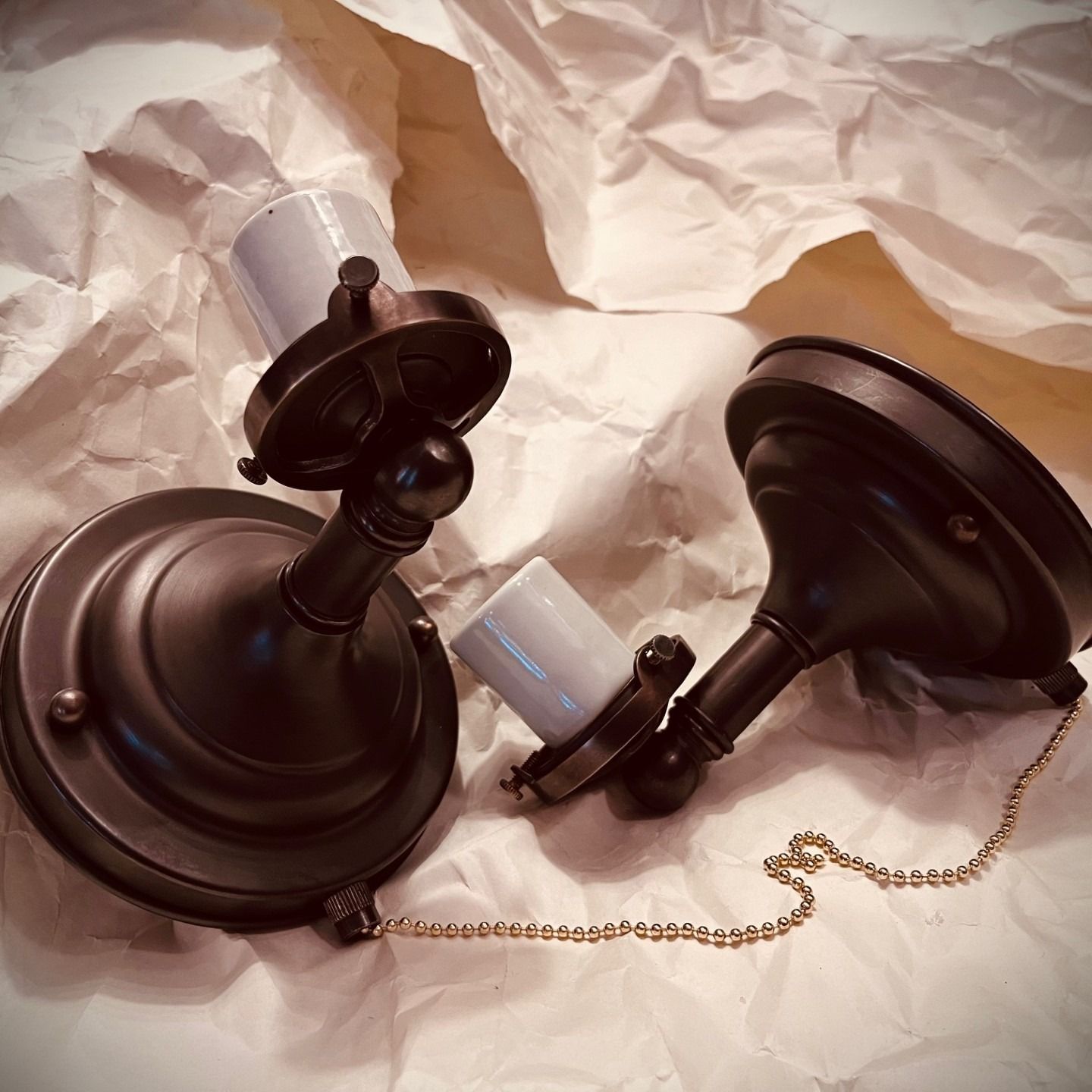 Two dark bronze-colored light fixtures with pull chains rest on crumpled paper.