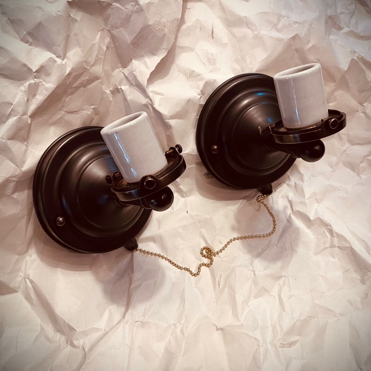 Two black, antique-style light fixtures with white ceramic sockets, connected by a gold chain, on wrinkled paper.
