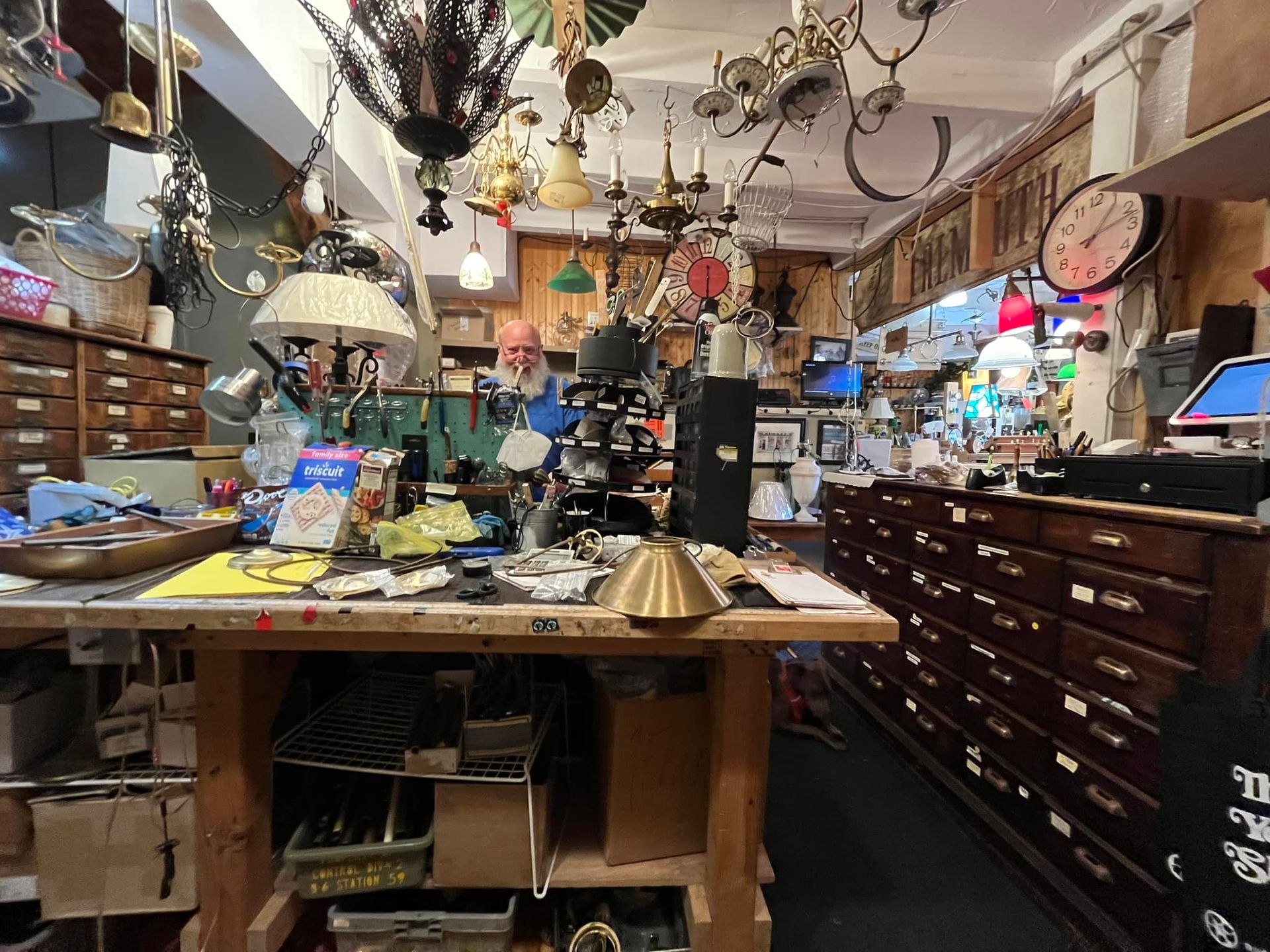 A cluttered shop interior filled with lamps and parts. A person stands behind a work table.