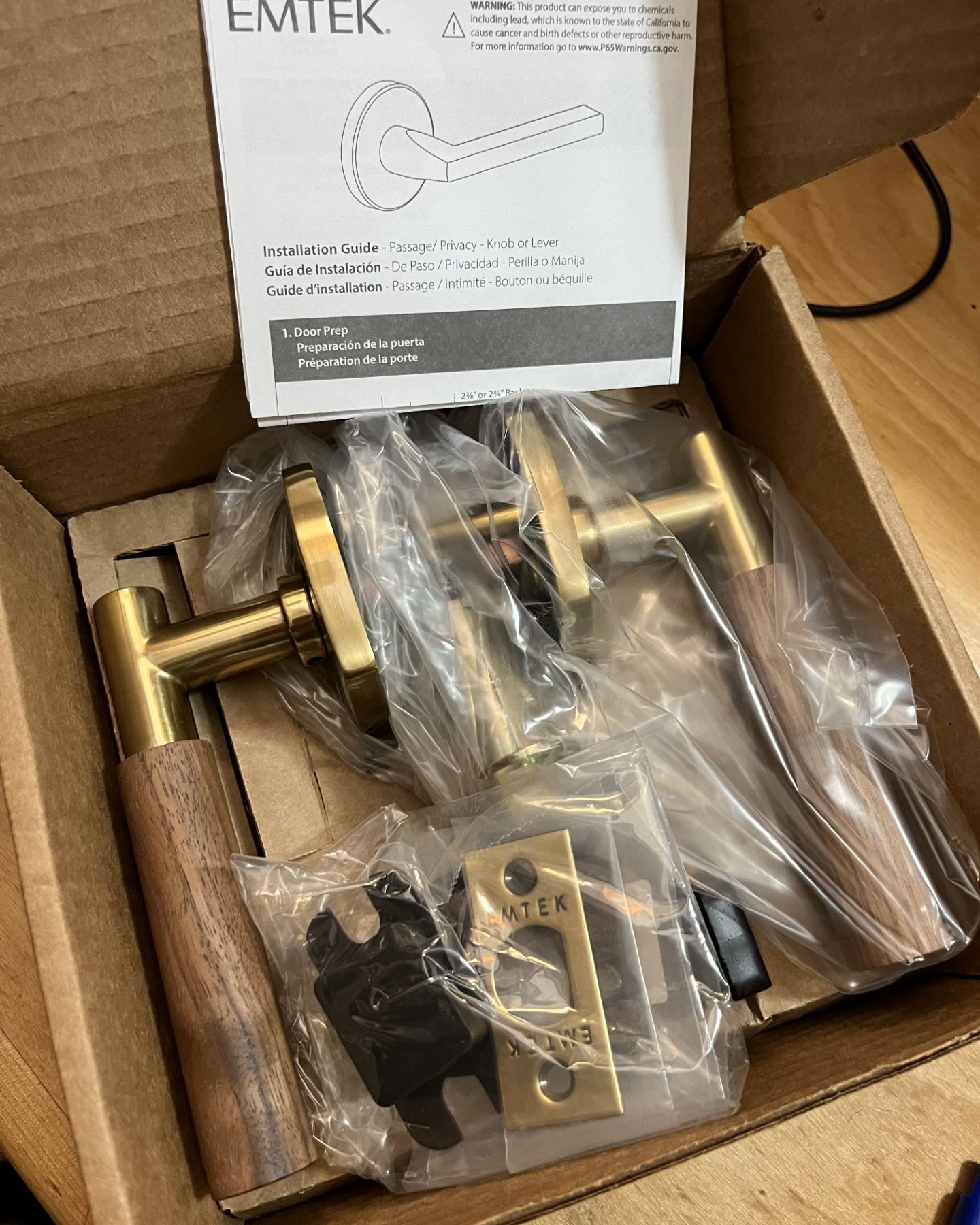 Box of new door hardware, featuring brass and wood lever handles, a strike plate, and instructions.
