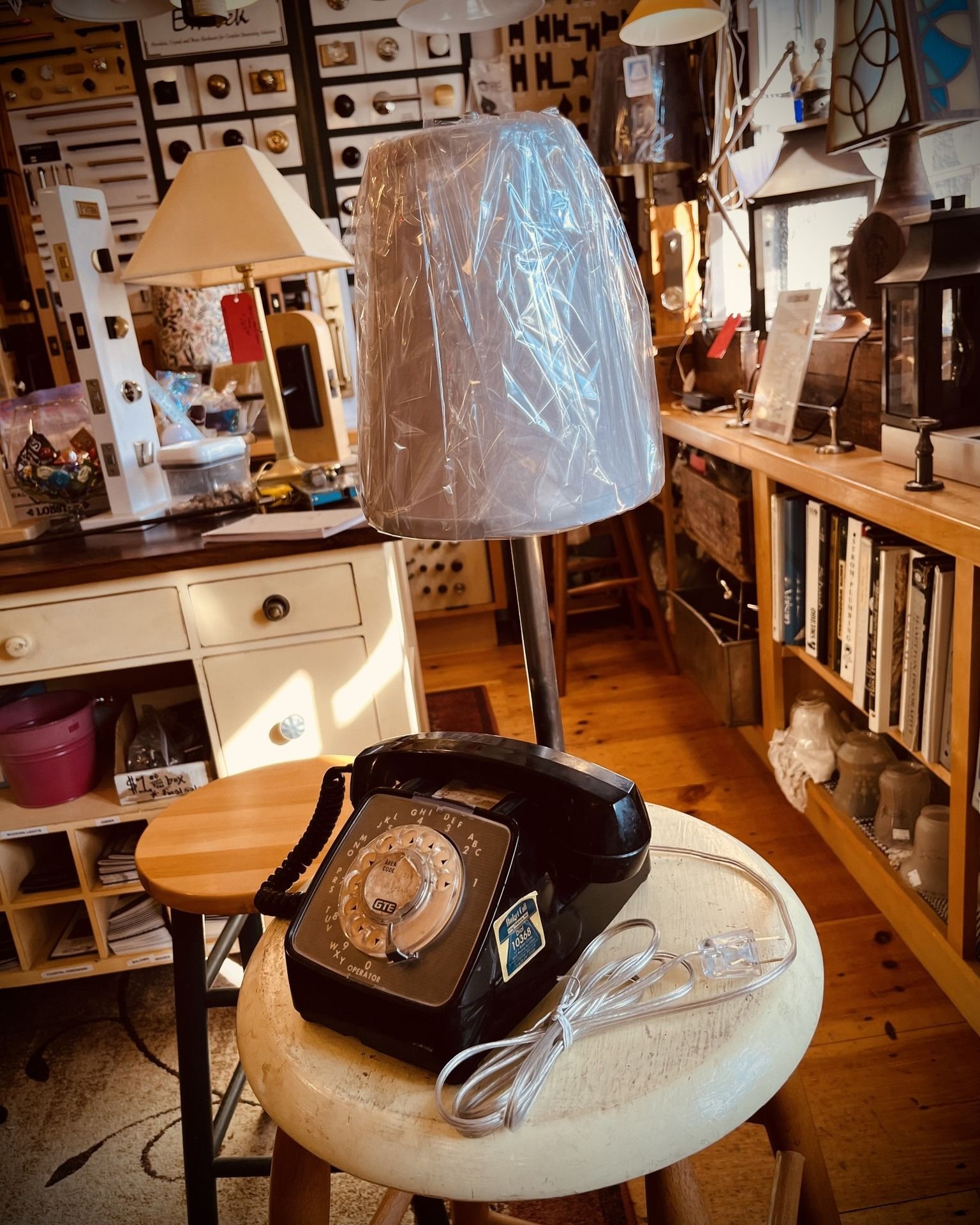 Black rotary phone on a stool, topped with a lamp with a plastic-covered shade in a cluttered shop.