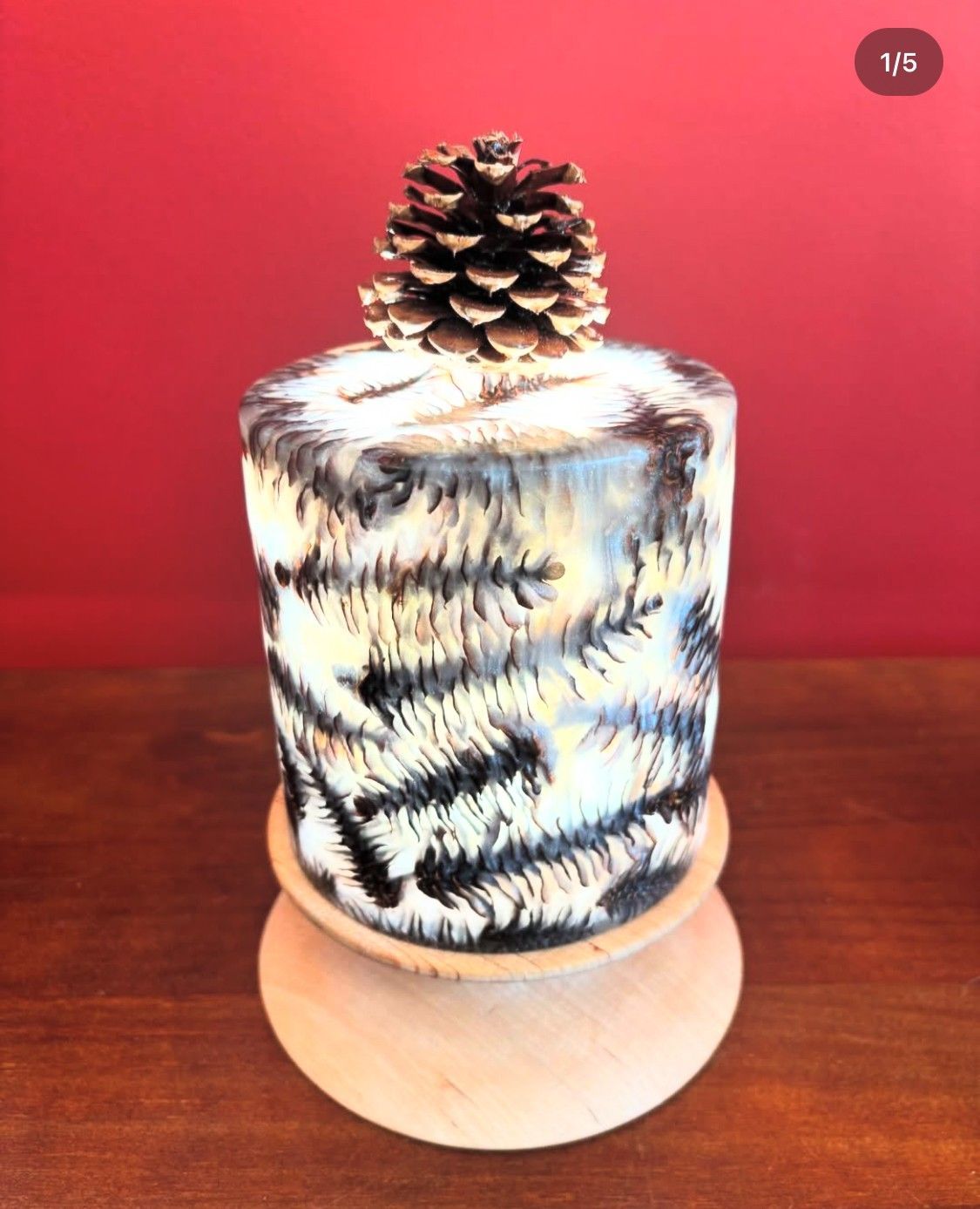 Cylindrical art piece with pine needle design, topped with pinecone, on a wooden base.