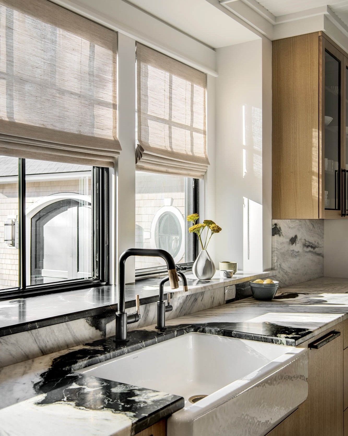 Kitchen with windows, a white sink, and black and white countertop. Neutral color window shades and wood cabinets.