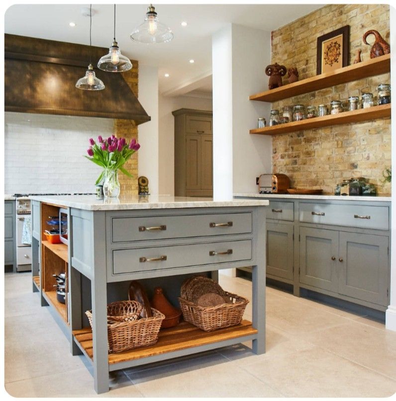 Gray kitchen island with marble countertop, open shelves, and wooden accents, next to brick wall with floating shelves.