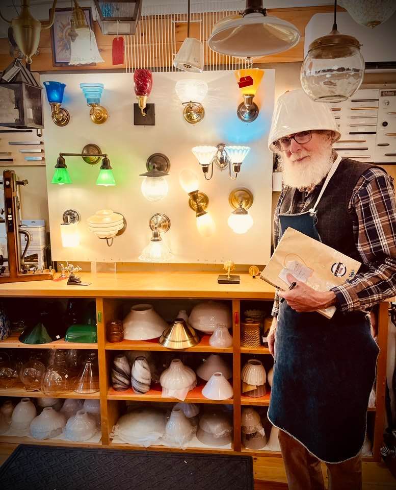 Man in shop poses next to wall display of light fixtures. He wears a lampshade on his head.