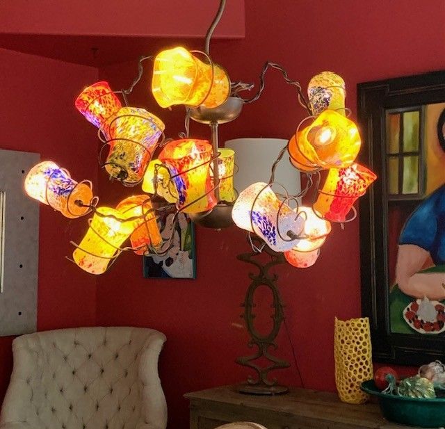 Chandelier with colorful, glowing lamps against a red wall. Interior setting with artwork and furniture.