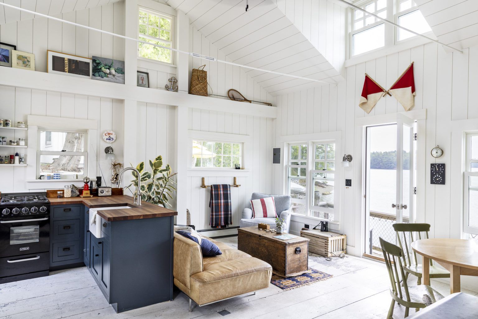 Bright, white-painted interior of a cozy cabin. Kitchen with a blue island and a living area with water views.
