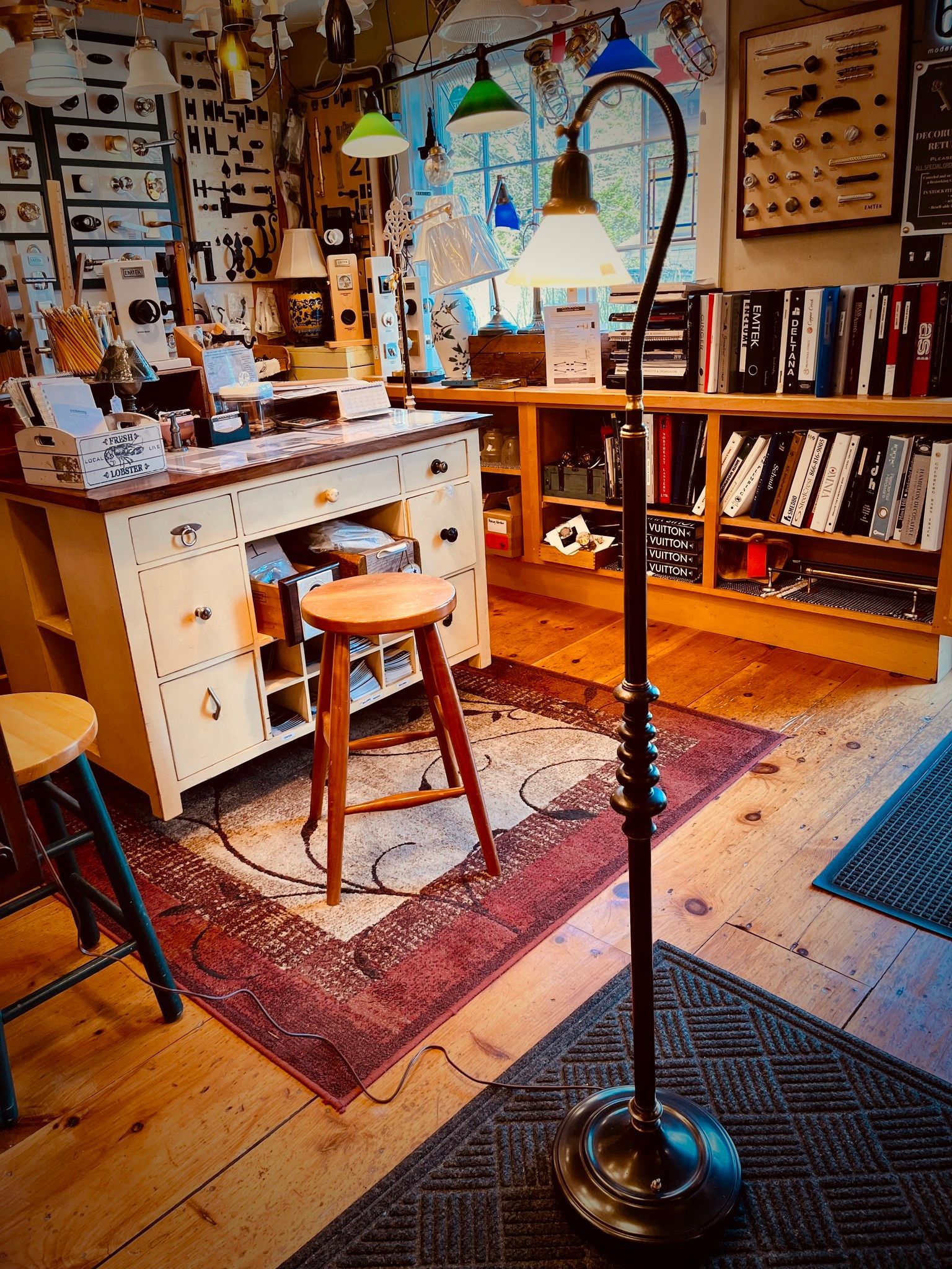 Workshop interior with a standing lamp, work table, stool, rug, and shelves filled with items.
