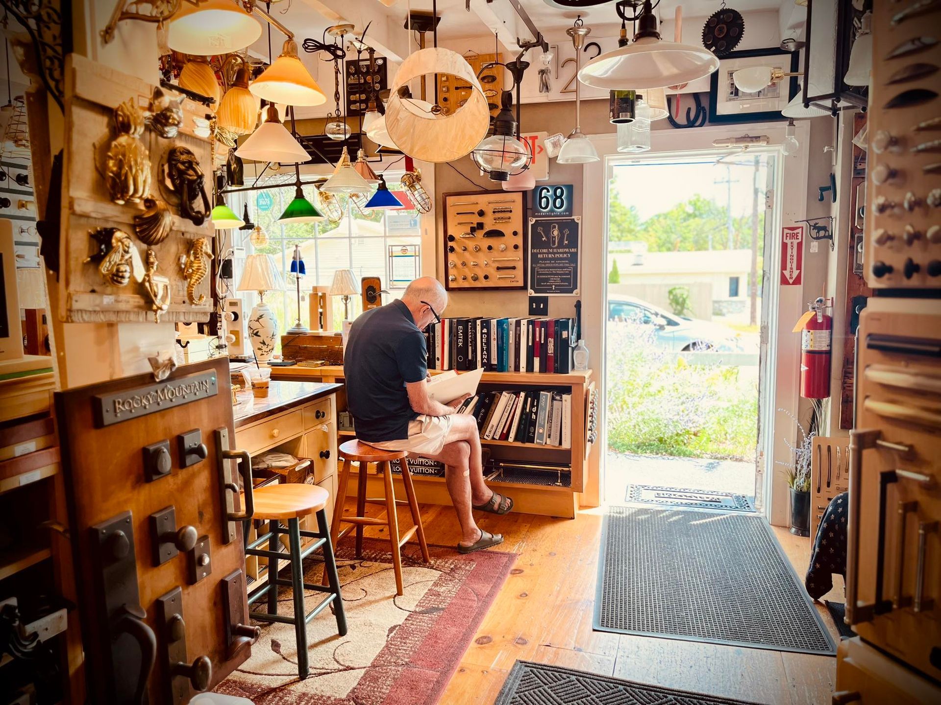 A man sits in a cluttered shop with many lamps, reading a book. Shop doorway leads outside.