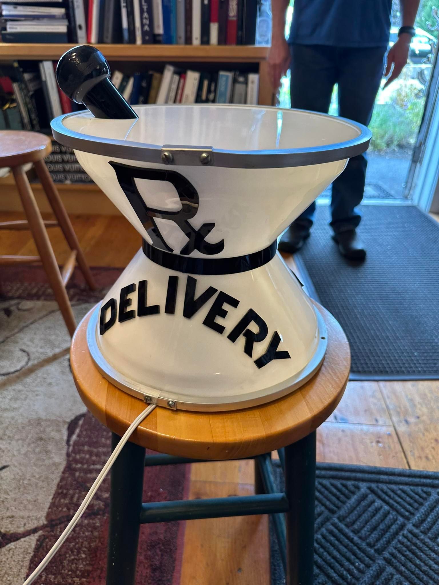 White pharmacy delivery sign with a mortar and pestle on a stool. A person stands nearby.