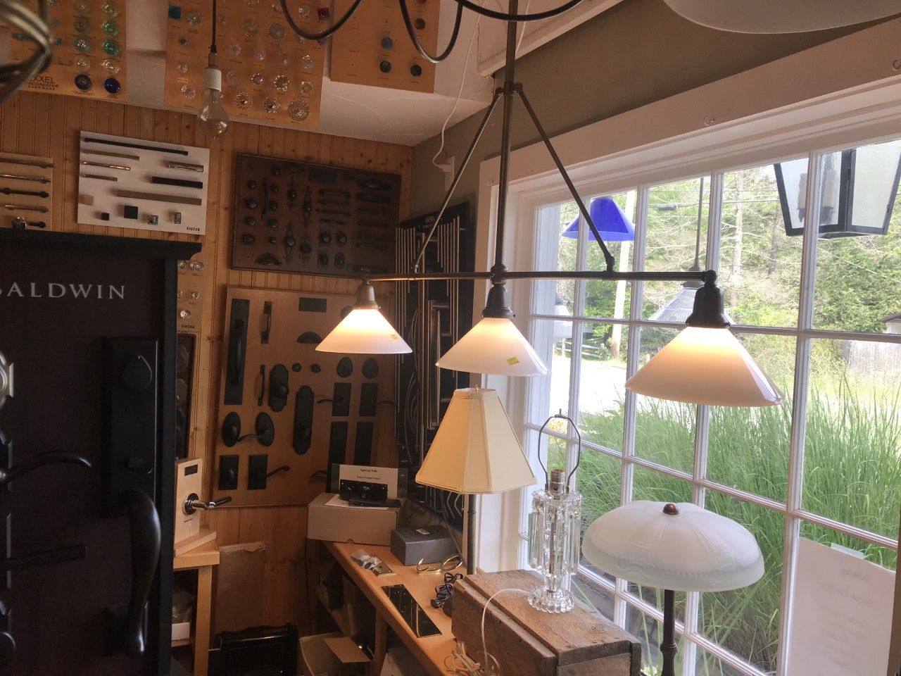 A display room with light fixtures. A three-bulb pendant lamp hangs by a window. Shelves hold more lamps and hardware.