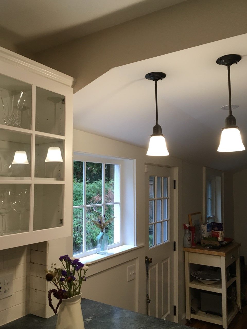 Kitchen with glass-fronted cabinets, arched doorway, two pendant lights, and a window overlooking greenery.