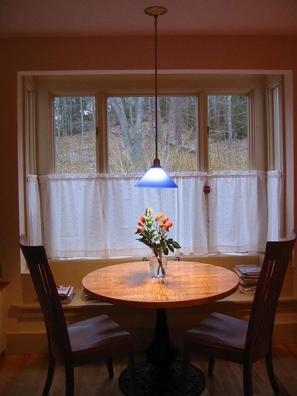 A small, round table set for two with a flower bouquet, beneath a blue-tinted light, by a window with white curtains.