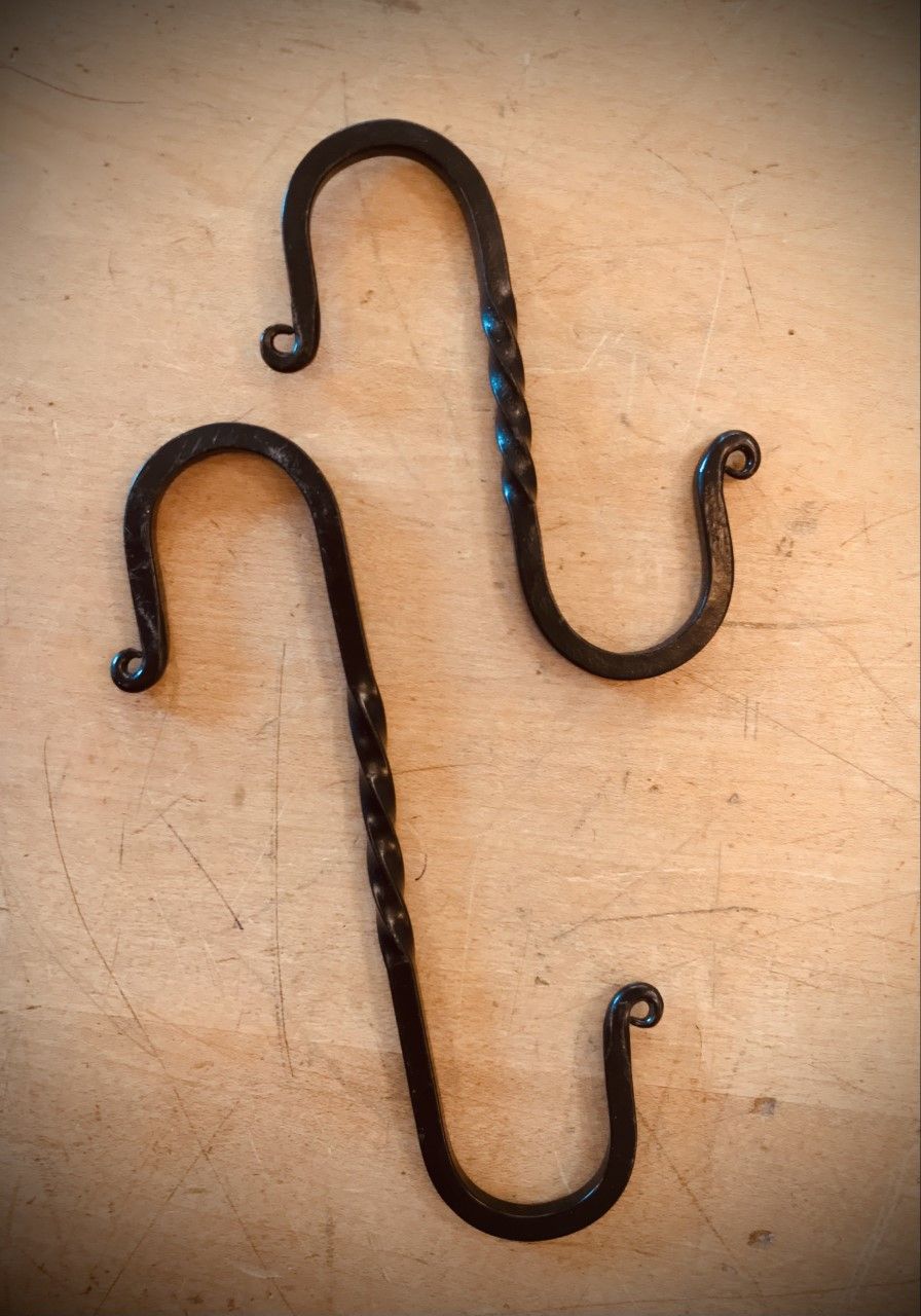 Two black, wrought-iron S-hooks on a light-brown wooden surface, one with a twisted center.