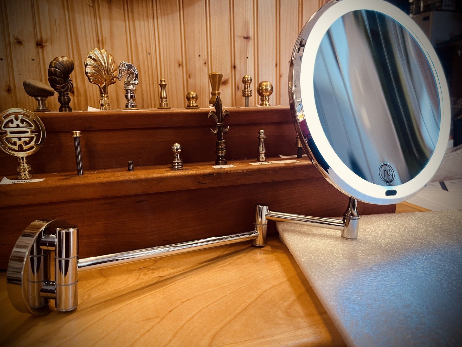 Chrome magnifying mirror with LED lights, on wooden surface. Background displays various metallic sculptures.