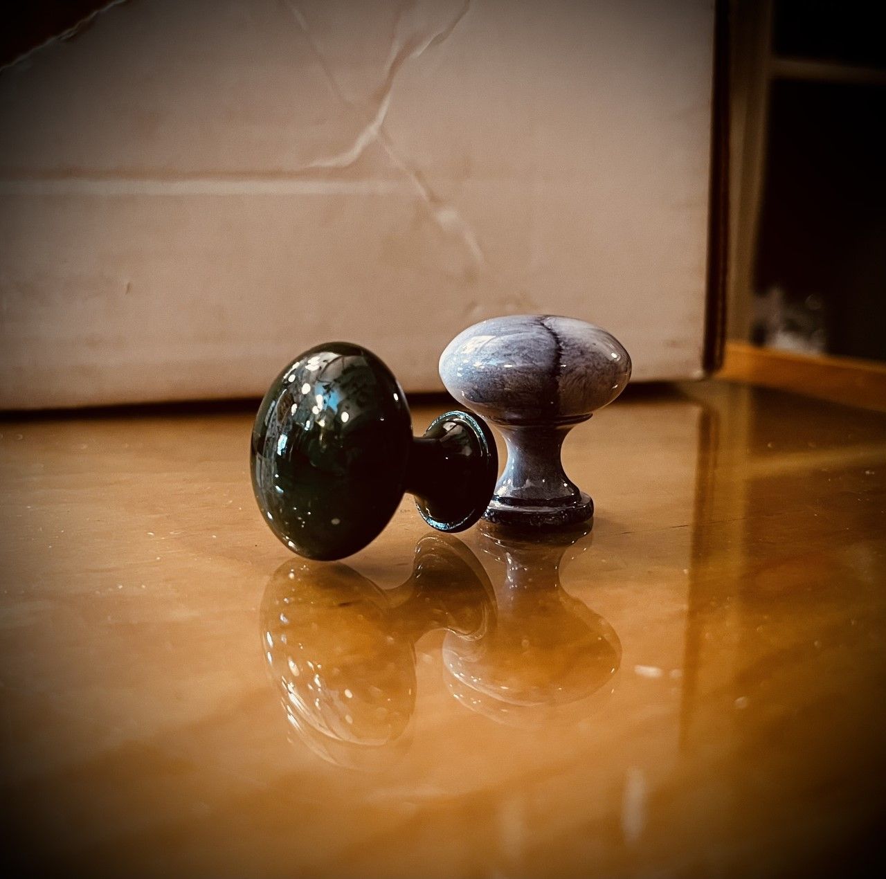 Two decorative cabinet knobs, one glossy green, one matte gray, on a reflective wooden surface.