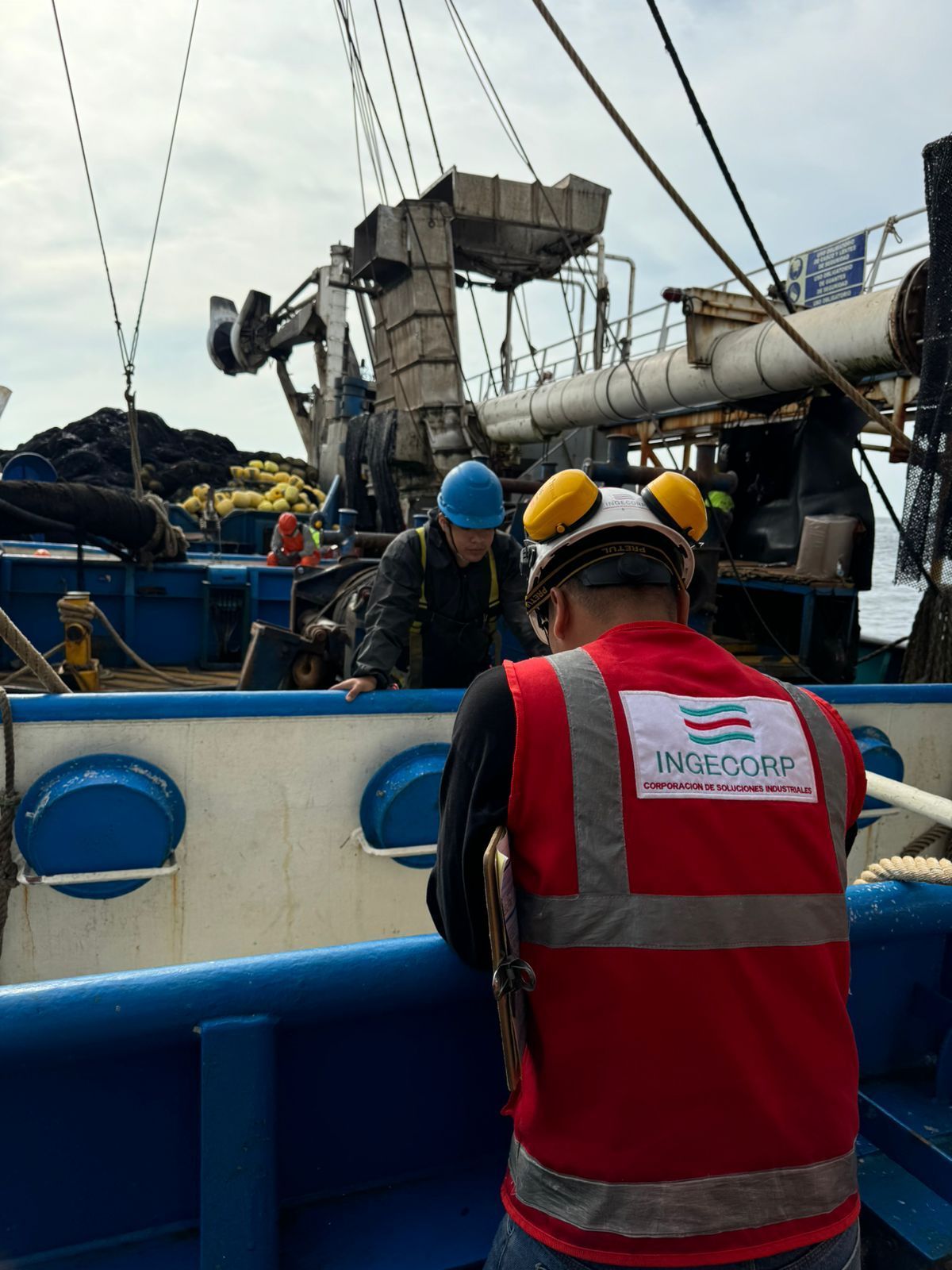 Trabajadores en un barco con equipo de seguridad. Uno inspecciona algo mientras otro permanece cerca.