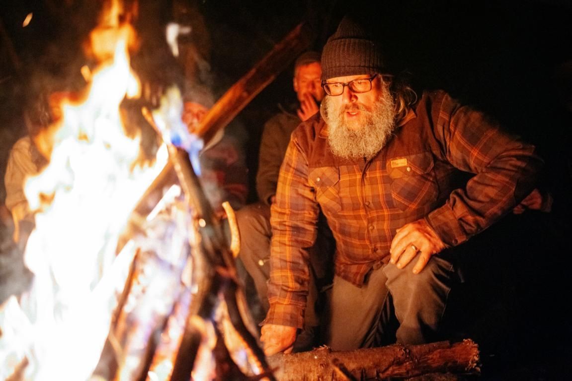 Ein Wanderer in einer neonfarbenen Jacke und mit einem großen Rucksack geht durch einen nebligen Wald auf andere Personen zu, die sich in der Nähe eines kleinen weißen Zeltes befinden.