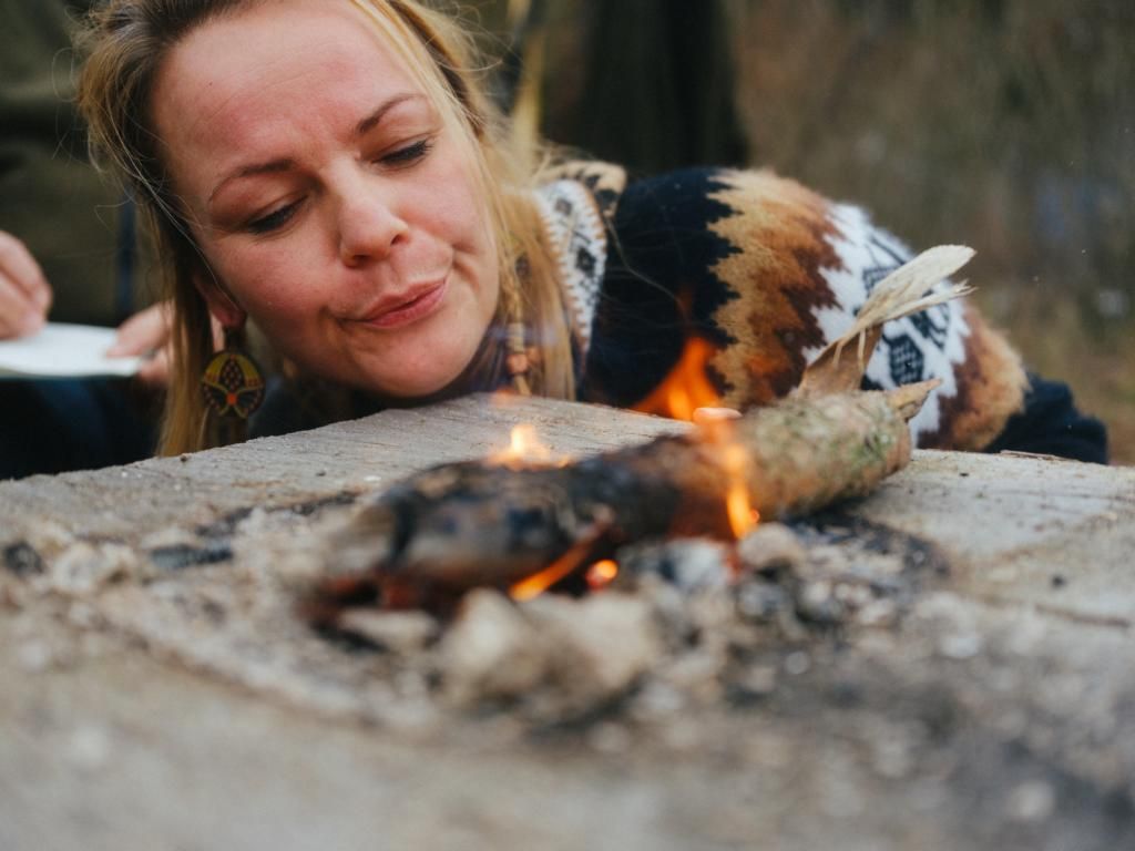 Eine bärtige Person in kariertem Hemd und Mütze hockt nachts an einem hell erleuchteten Lagerfeuer, im Hintergrund sitzen weitere Personen.
