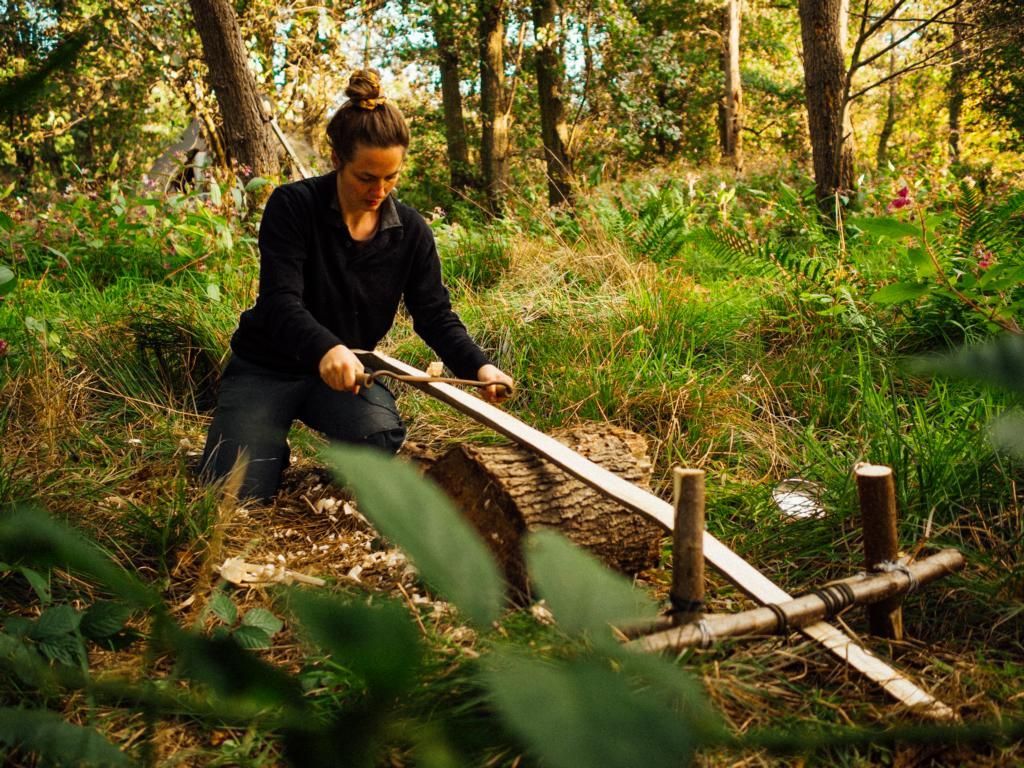 Eine Person kniet in einem Wald und sägt mit einer langen Handsäge einen von einem Holzrahmen gestützten Baumstamm durch.