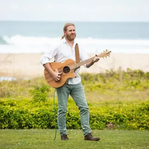 A Man is Standing in a Field Holding an Acoustic Guitar — Chop 'n Chill South West Rocks in South West Rocks, NSW