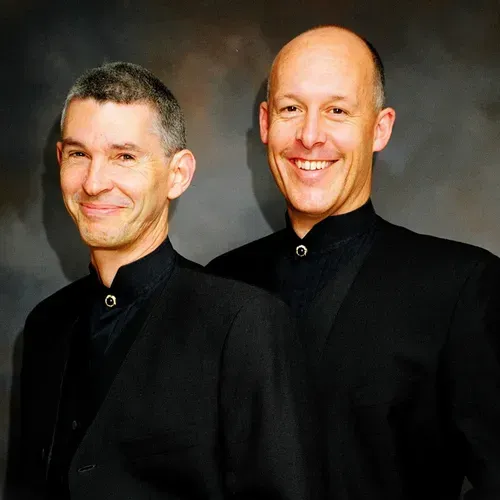 Two men in black suits are standing next to each other and smiling.