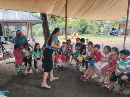 A woman is standing in front of a group of children sitting under a tent.