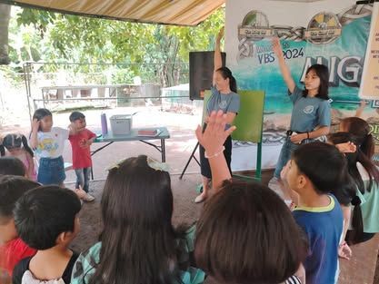 A woman is standing in front of a group of children raising their hands.
