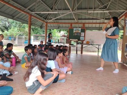 A woman is standing in front of a group of children sitting on the floor.