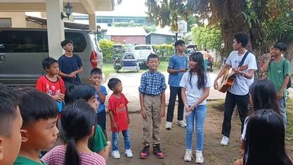 A group of children are standing around a man playing a guitar.