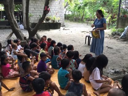 A woman is standing in front of a group of children sitting on the ground.
