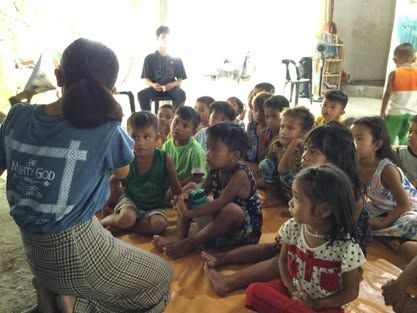 A woman is kneeling down in front of a group of children sitting on the floor.