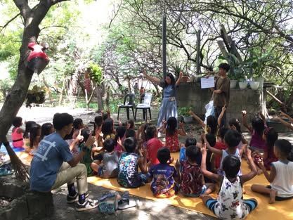 A group of children are sitting on a mat under a tree.