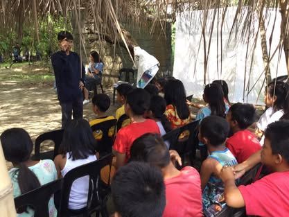 A man is standing in front of a group of children sitting in chairs.