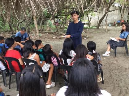 A boy is standing in front of a group of people sitting in chairs.