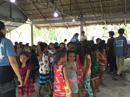 A group of children are standing in a line under a thatched roof.