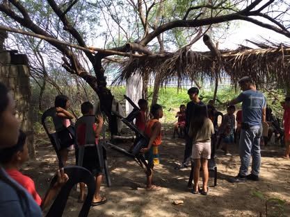 A group of children are standing in a circle under a thatched roof.