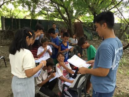 A group of children are sitting around a man holding a piece of paper.