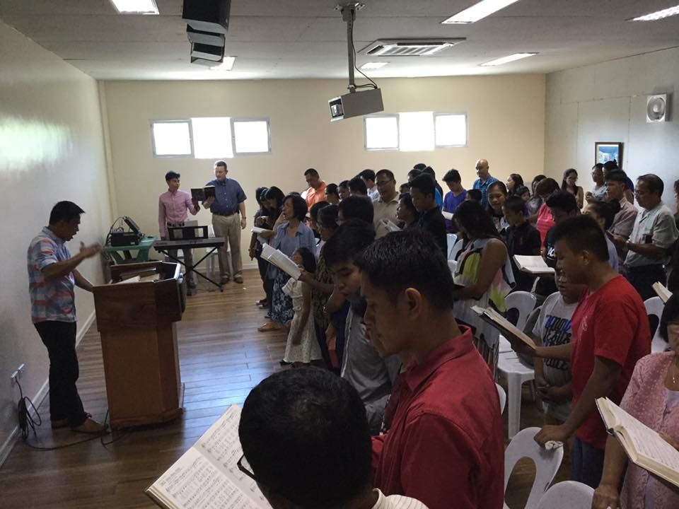 A group of people are singing in a room while a man stands at a podium.