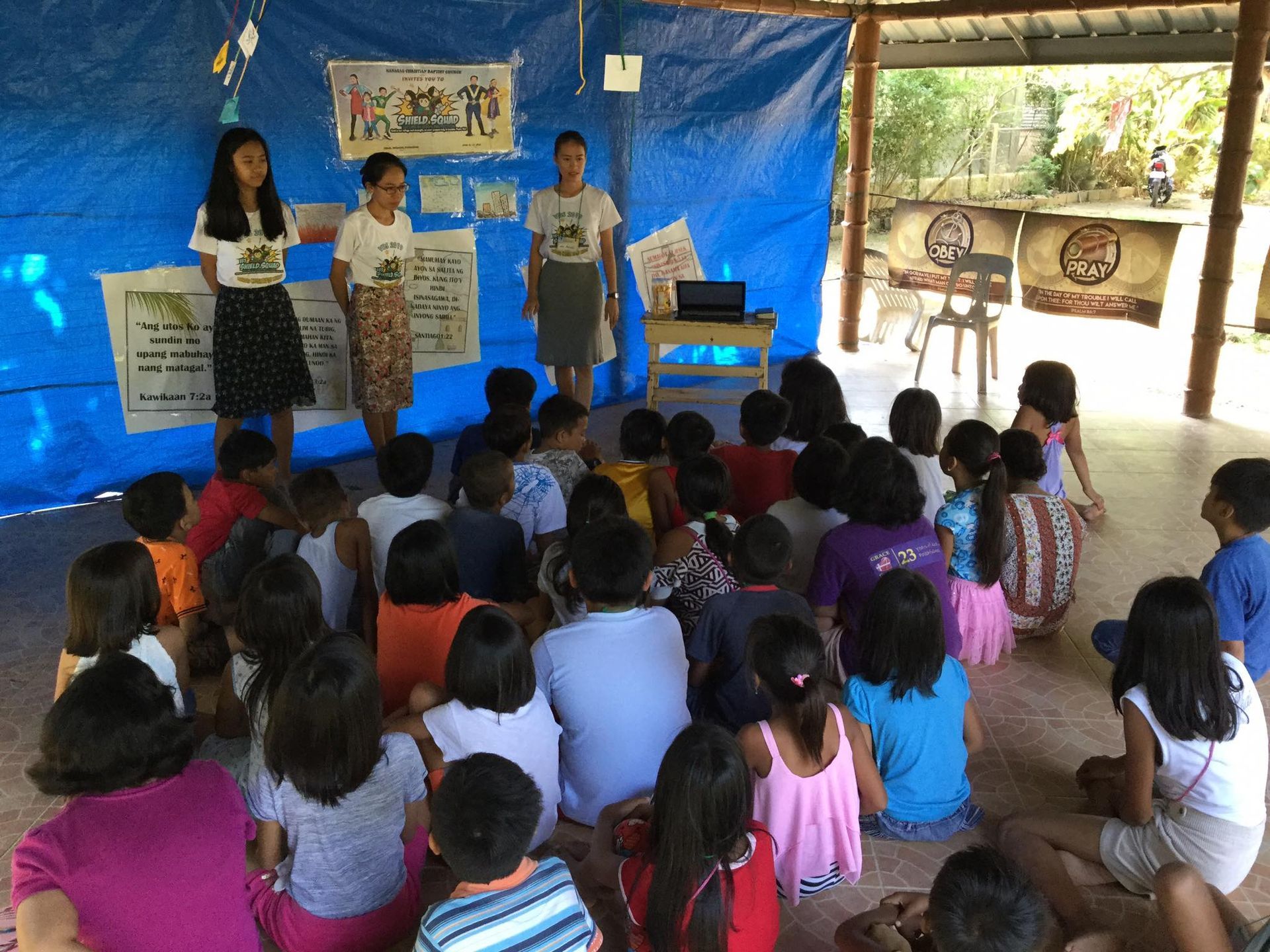 A group of children are sitting on the floor in front of a blue tarp.