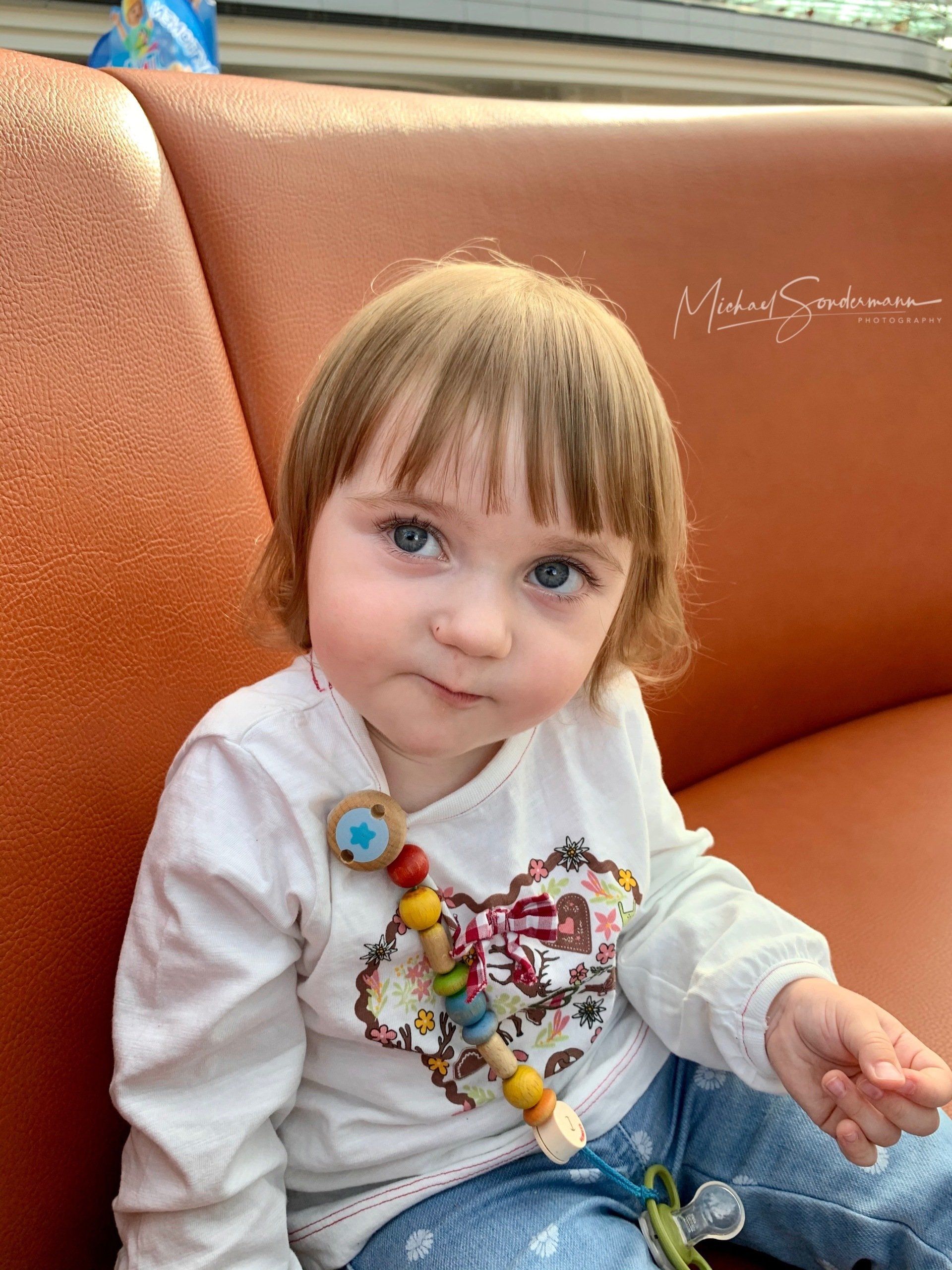 Ein junges Mädchen sitzt grinsend auf eine Bank und shakert mit der Kamera.