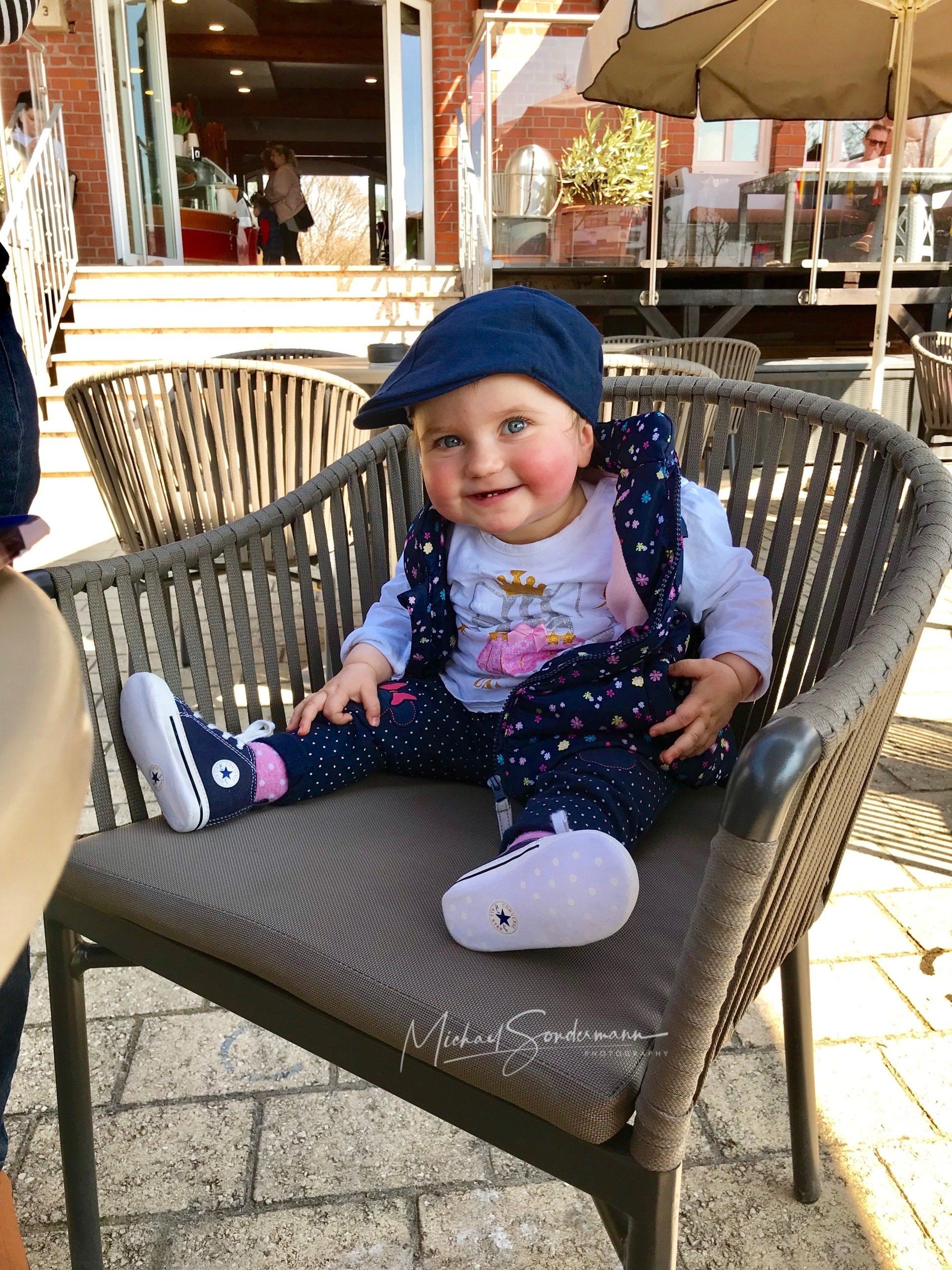 Ein junges Mädchen grinst in die Kamera und sitzt dabei auf einen Stuhl in einem Eiscafe im Sommer.