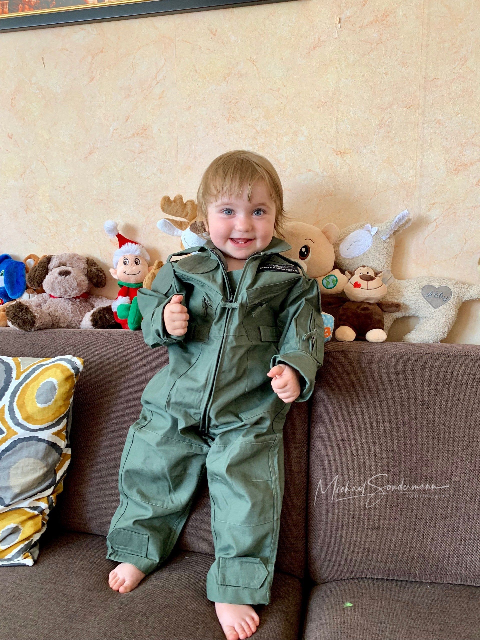 Ein junges Mädchen steht im Fliegeroverall und grinsend auf dem Sofa und hat im Hintergrund viele Kuscheltiere stehen.