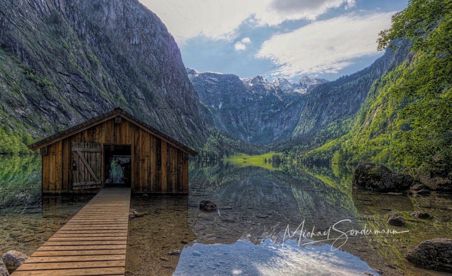 A wooden hut is sitting on a dock next to a lake surrounded by mountains.