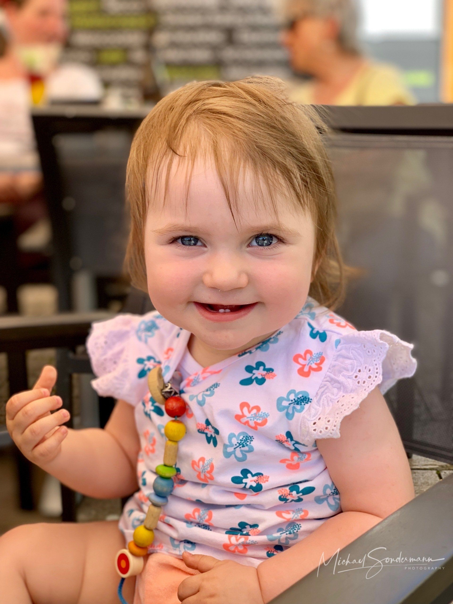 Ein junges Mädchen sitzt im Sonnenschein in einem Cafe auf dem Stuhl und lacht.