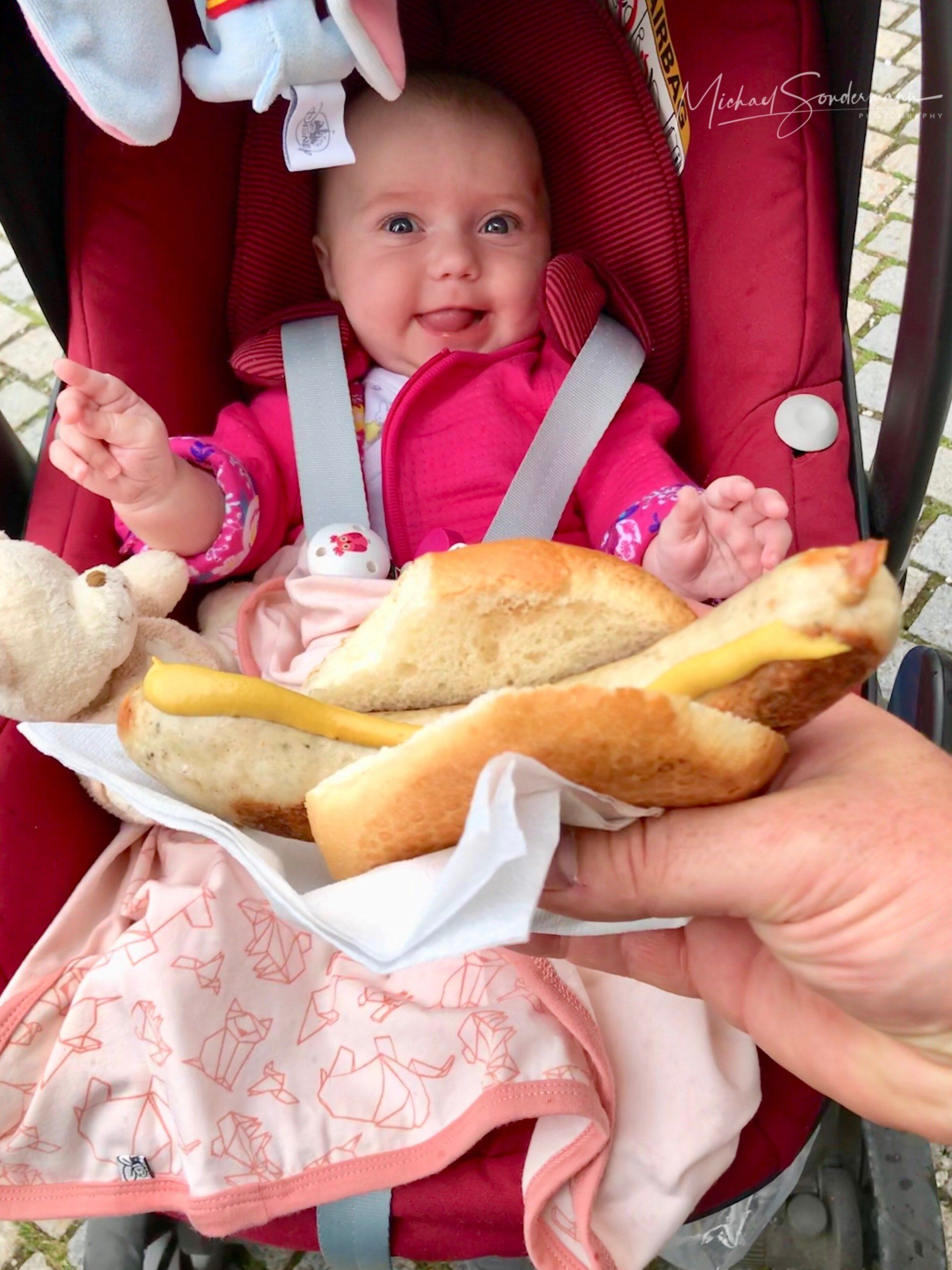 Ein junges Mädchen sitzt grinsend im Kinderwagen und hält vor sich eine Bratwurst im Brötchen.