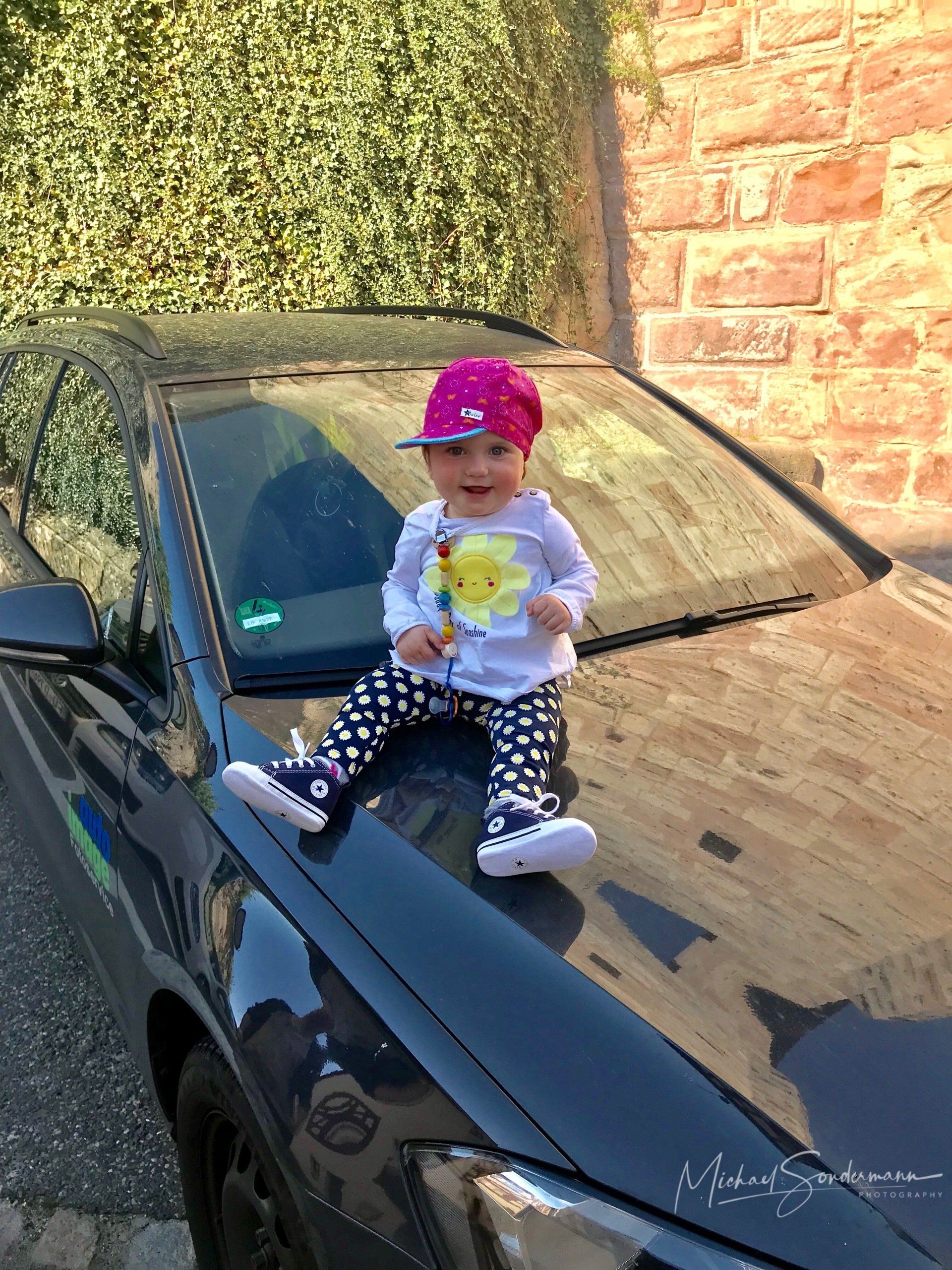 Ein junges Mädchen sitzt grinsend mit einen Capi auf dem Kopf auf der Motorhaube eines schwarzen Auto.