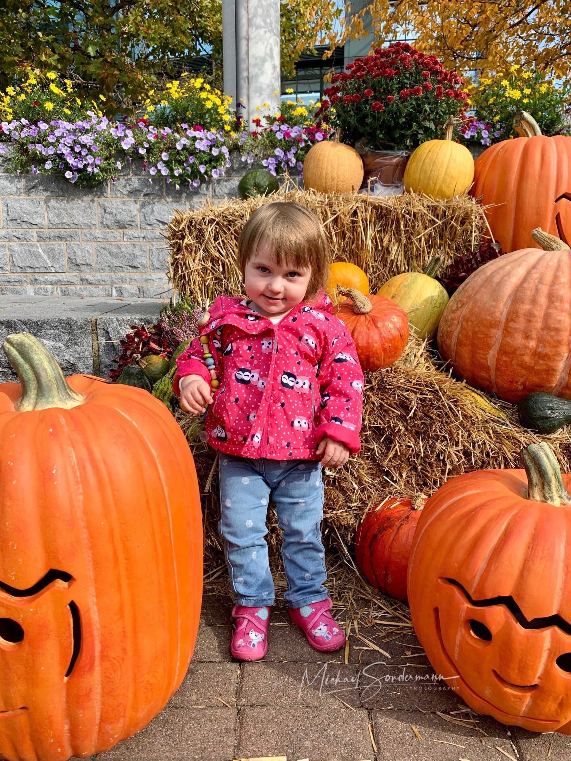 Ein junges Mädchen steht lächelnd zwischen mehreren Strohballen und Kürbissen